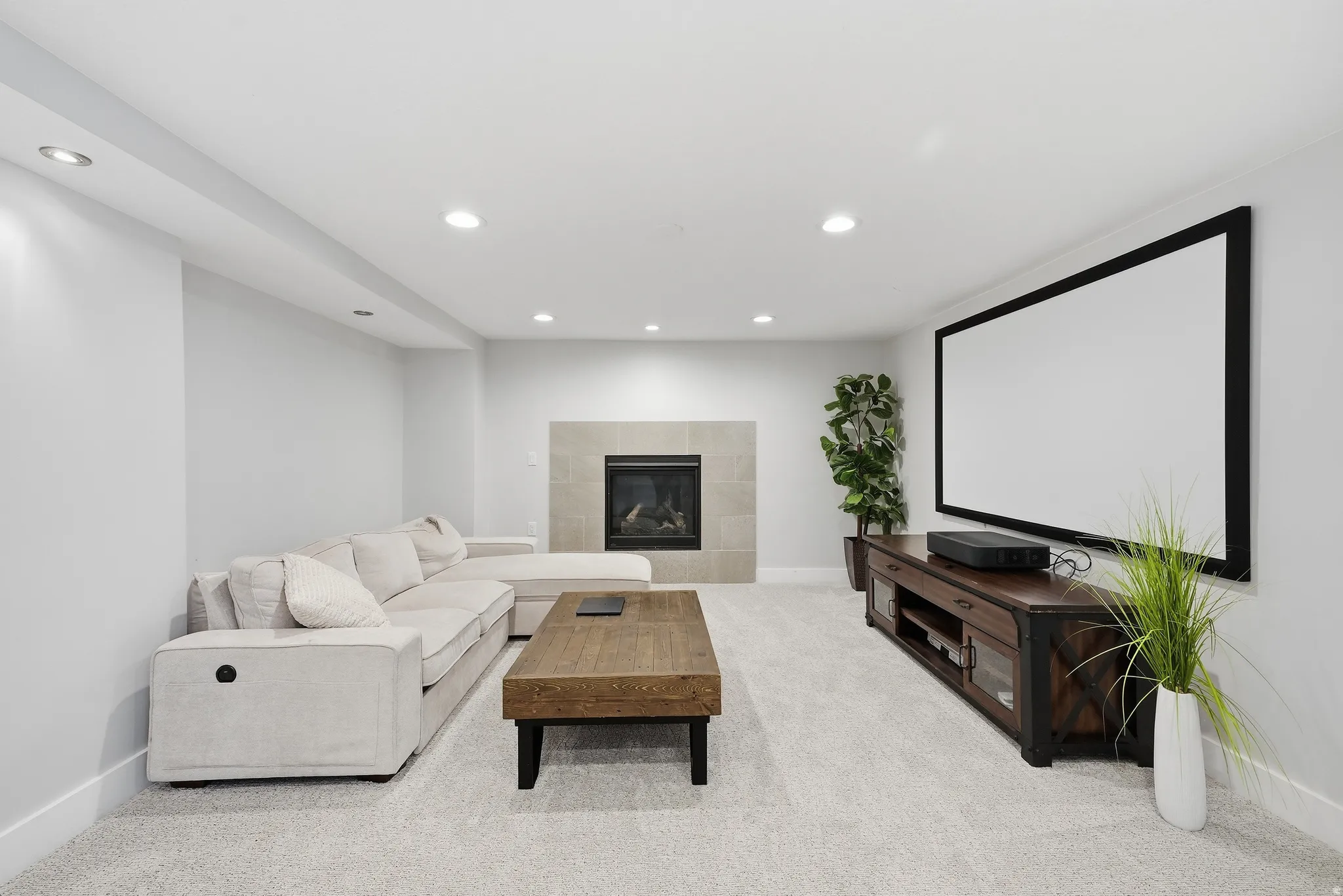 Luxurious basement recreation room featuring recessed ceiling lighting, a wood-finish entertainment center with built-in media storage, a gas fireplace with stone surround, light-finish flooring throughout, and a large projection screen wall ideal for ent