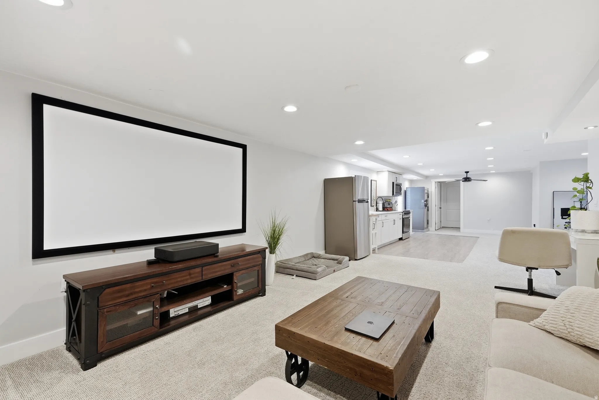 Recessed lighting, large projection screen, wood-finish media console, light-toned carpeting, and a kitchen area with stainless steel appliances