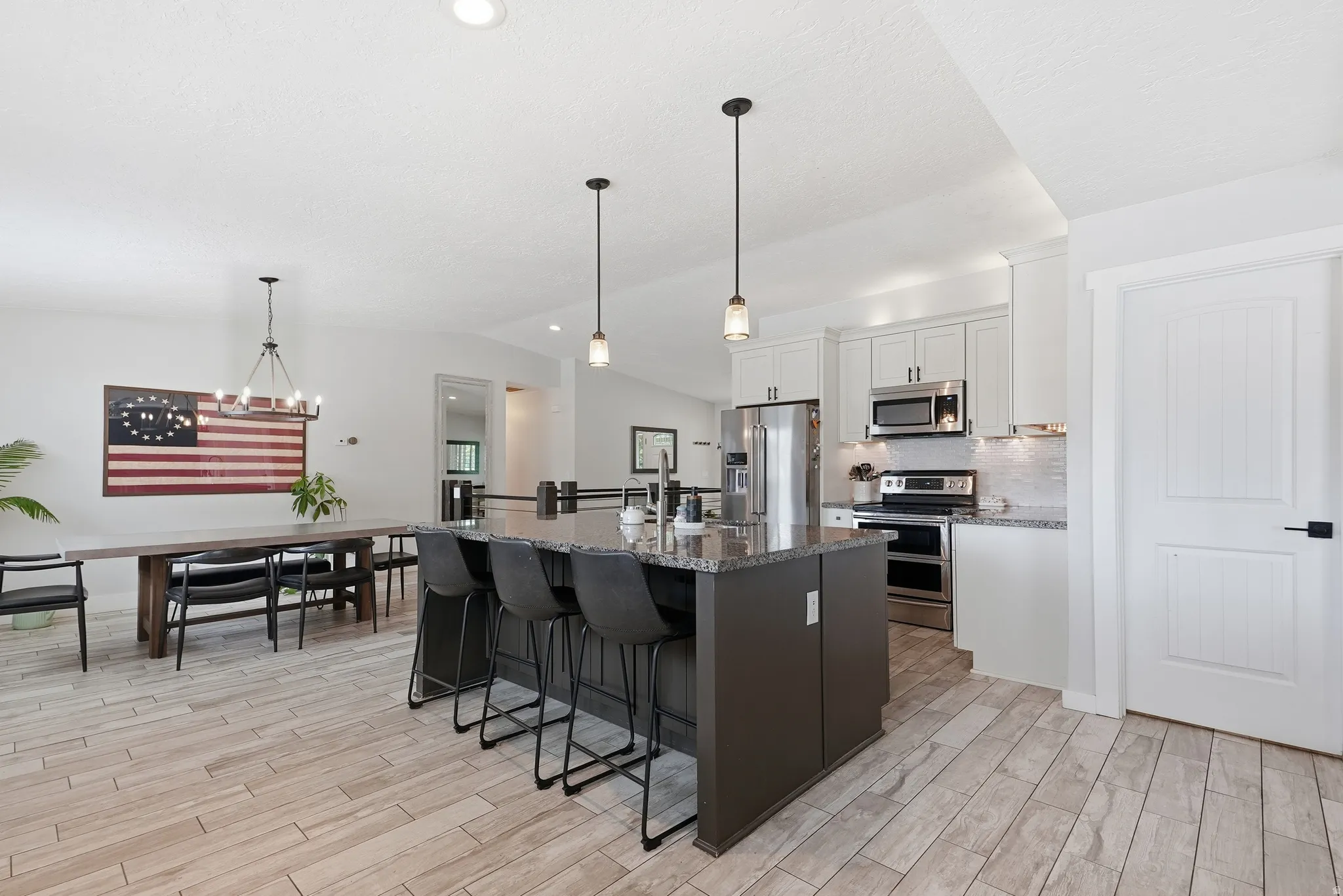 Open-concept kitchen and dining area featuring stainless steel appliances, dark cabinetry with waterfall-edge island seating for multiple guests, pendant lighting, wood-finish flooring throughout, and abundant natural light from recessed ceiling details
