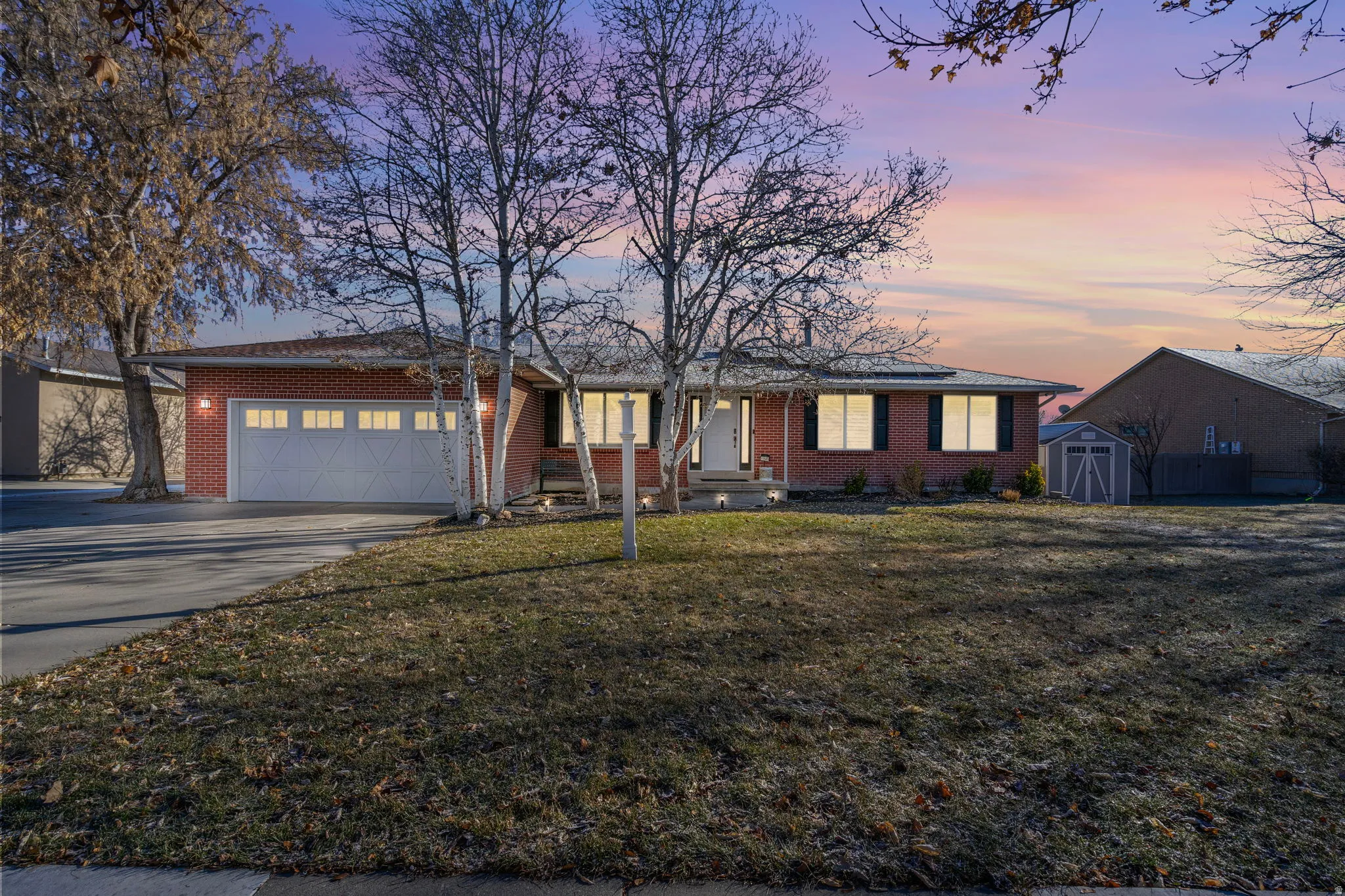 Brick exterior with attached garage, paved driveway, mature trees, and a storage shed