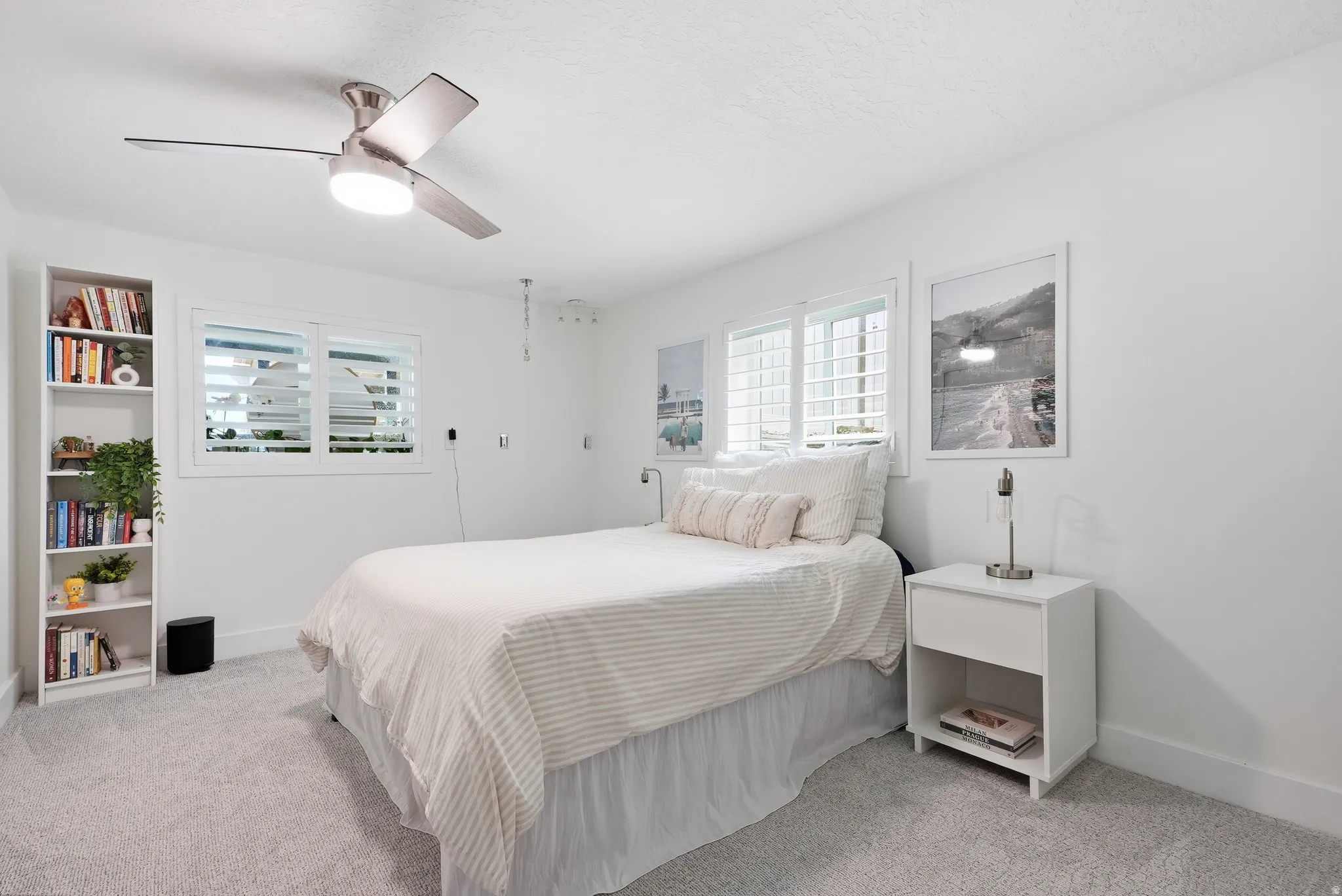Bright, light-filled bedroom featuring carpet flooring, a ceiling fan with integrated lighting, built-in shelving with display storage, and shuttered windows allowing natural light