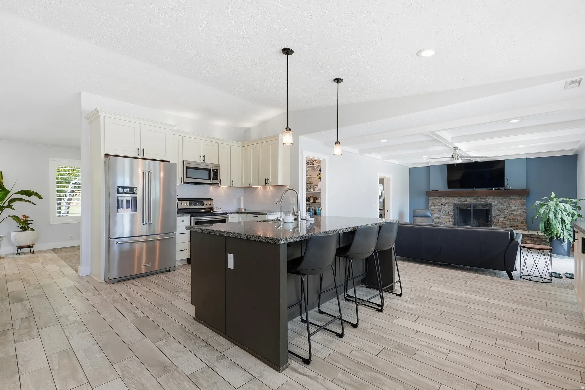 Open-concept kitchen and living area featuring stainless steel appliances, a large center island with seating for multiple guests, wood-finish flooring throughout, pendant lighting, and abundant natural light from multiple windows