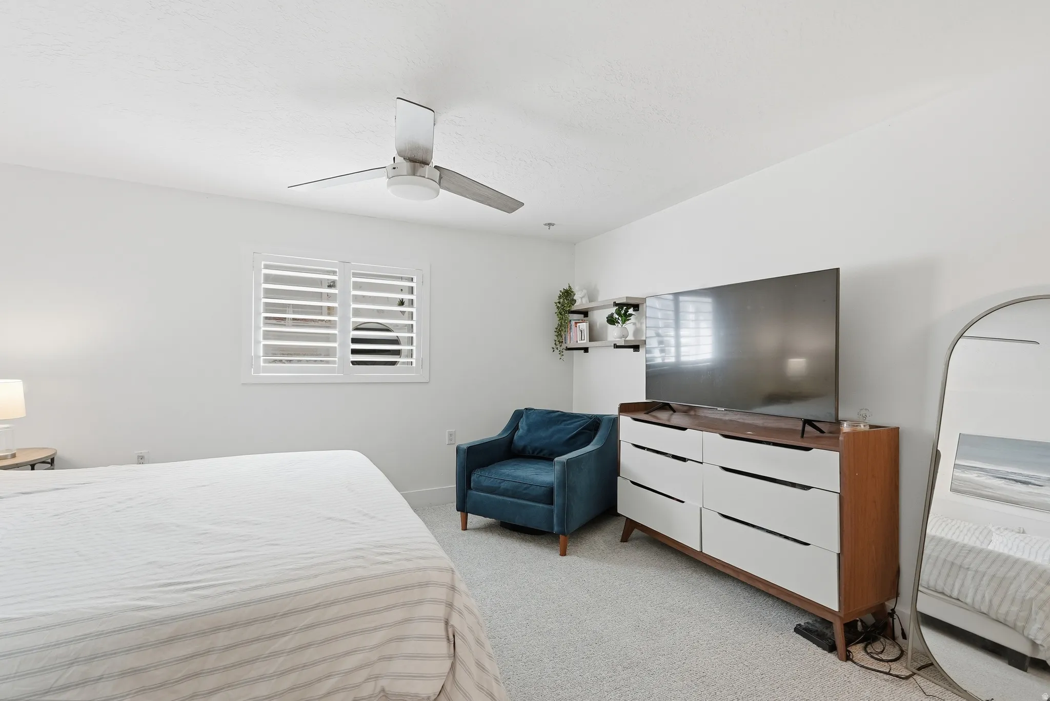 Bright secondary bedroom featuring neutral carpeting, ceiling fan with integrated lighting, shuttered window with natural light, and a wood-finish dresser with white cabinetry
