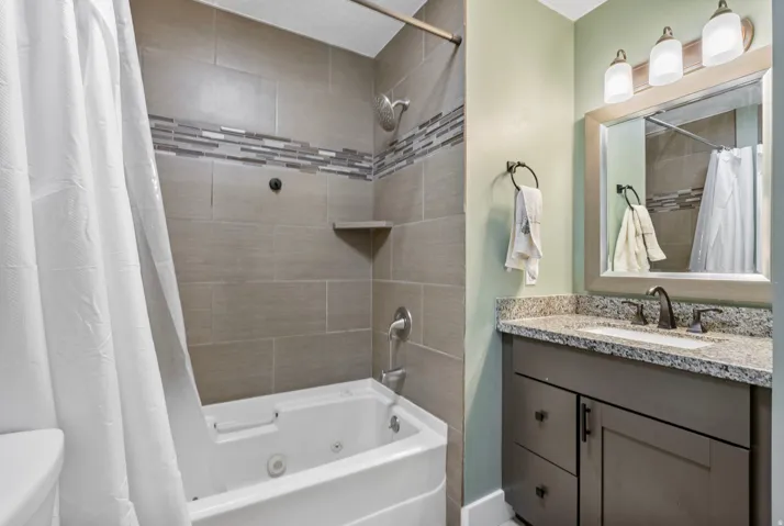 Bathroom featuring a built-in jetted tub, tiled shower surround with accent band, vanity with stone countertop, and an illuminated mirror