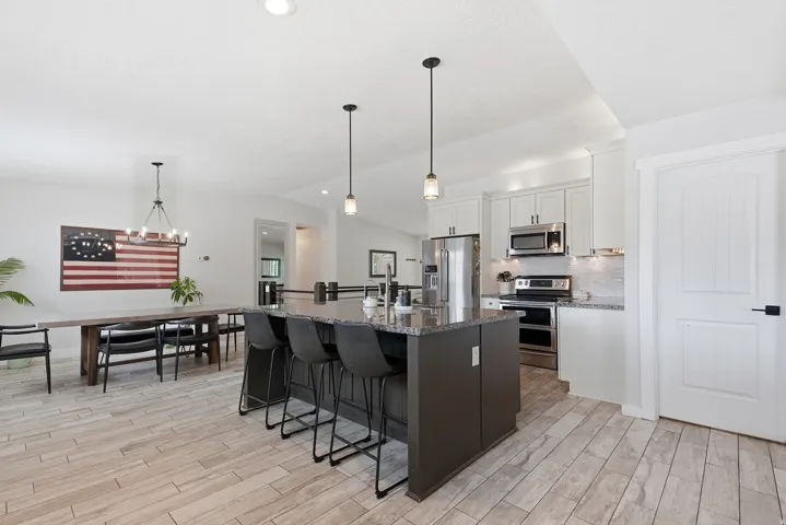 Open-concept kitchen and dining area featuring stainless steel appliances, dark cabinetry with waterfall-edge island seating for multiple guests, pendant lighting, wood-finish flooring throughout, and abundant natural light from recessed ceiling details