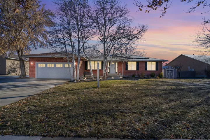 Brick exterior with attached garage, paved driveway, mature trees, and a storage shed