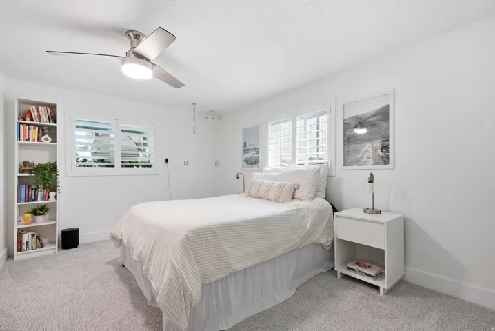 Bright, light-filled bedroom featuring carpet flooring, a ceiling fan with integrated lighting, built-in shelving with display storage, and shuttered windows allowing natural light