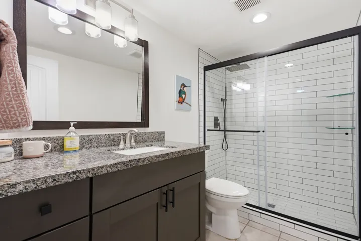 Spa-inspired ensuite bathroom featuring a dual-sink vanity with speckled stone countertop, framed mirror with integrated lighting, and a spacious walk-in shower with white subway tile surround and frameless glass enclosure