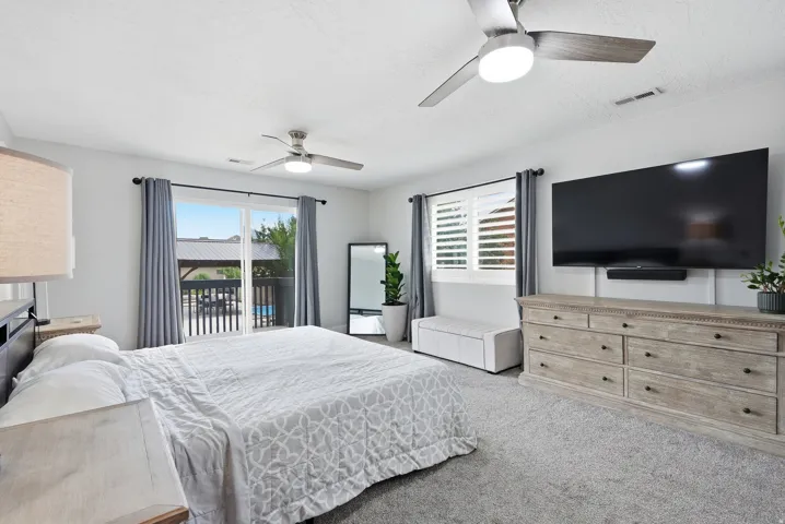 Sliding glass door leading to a private balcony, two ceiling fans with integrated lighting, window with plantation shutters, and light-toned carpeting