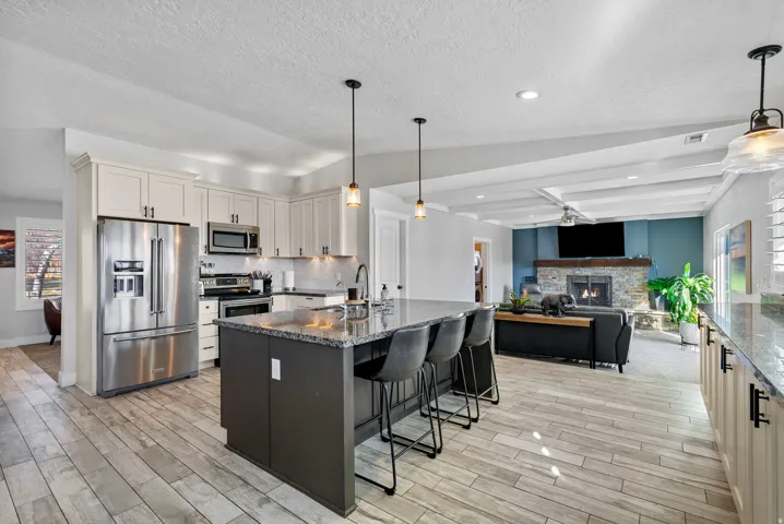 Expansive open-concept design with wood-finish flooring, a kitchen island with seating, a stone-faced fireplace, and exposed ceiling beams
