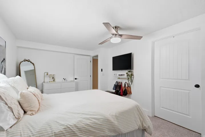 Bright secondary bedroom featuring neutral striped bedding, ceiling fan with brushed nickel finish, white six-panel closet doors, and built-in wall cabinetry with integrated television mounting and storage below