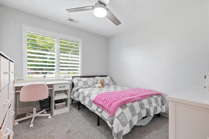 Bright secondary bedroom featuring a ceiling fan with integrated lighting, large multi-pane shuttered windows with natural light, neutral carpet flooring, and a built-in desk with storage shelving ideal for a workspace or study area