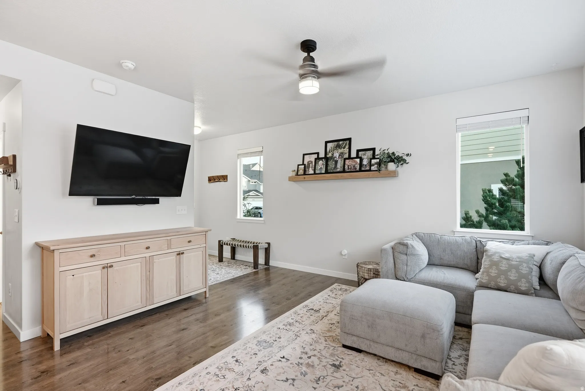Bright living room featuring hardwood flooring, a wall-mounted flat-screen television, light wood cabinetry with storage, ceiling fan with integrated lighting, and large windows allowing natural light throughout the space