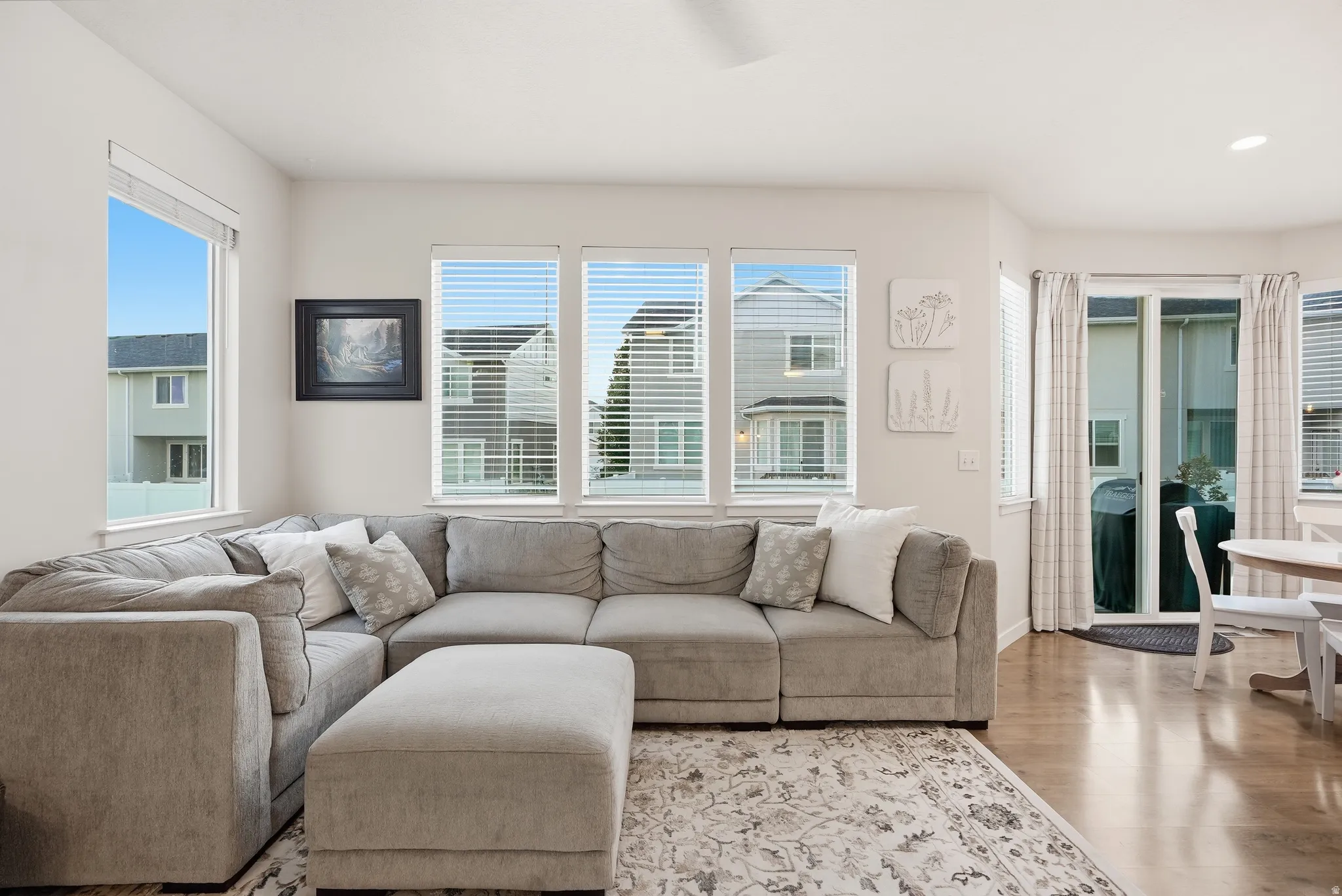 Bright, light-filled living room showcasing a sectional sofa arrangement, wood-finish flooring, and expansive multi-pane windows with operable shutters