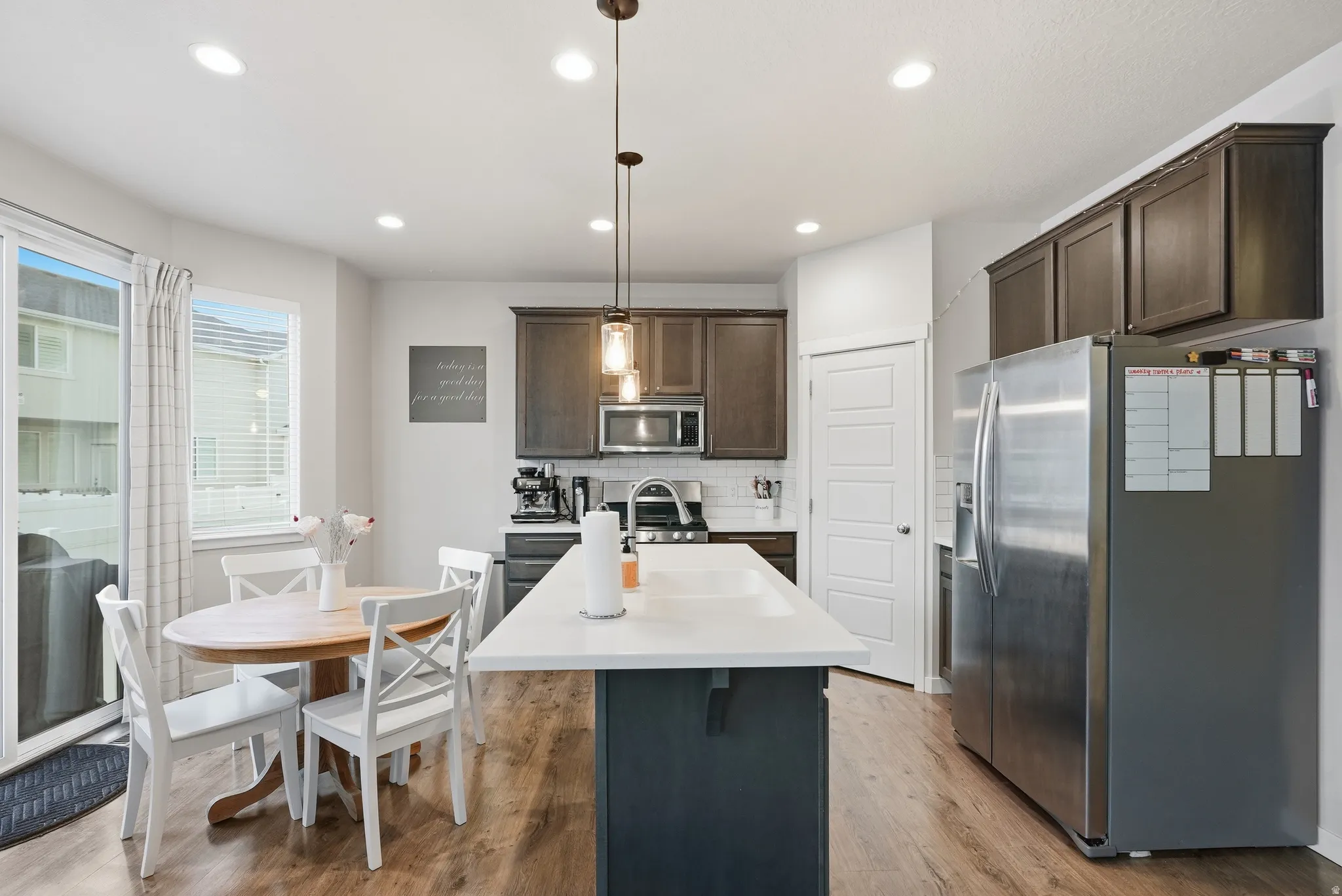 Modern kitchen featuring stainless steel appliances, a white quartz-topped island with seating, dark cabinetry with integrated hardware, wood-finish flooring, and recessed ceiling lighting throughout the open-concept space