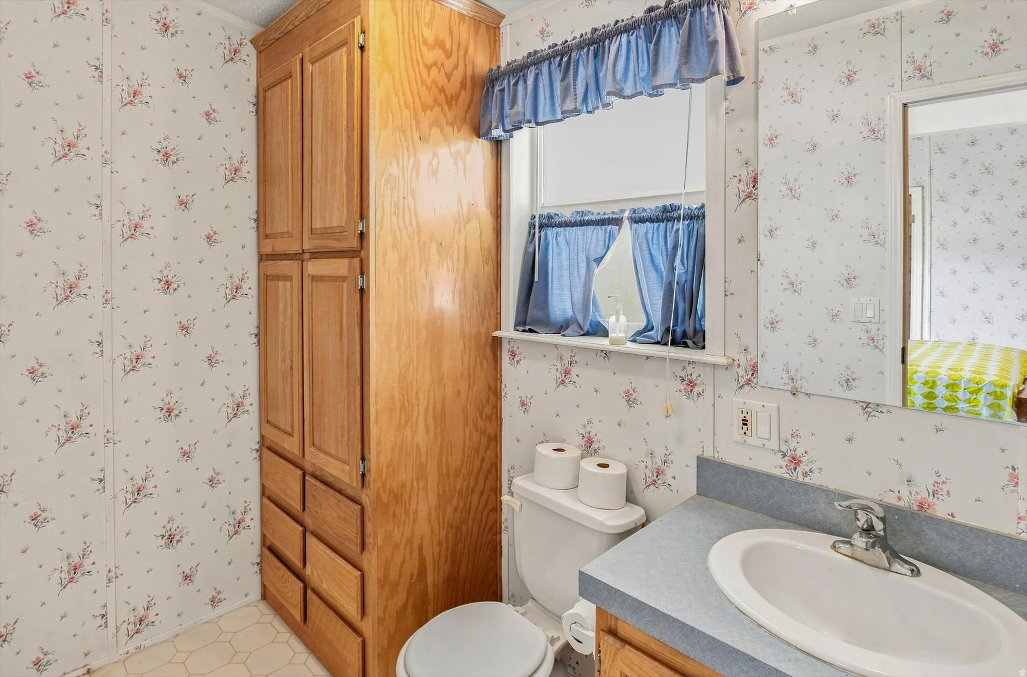 Full bathroom featuring a dual-sink vanity with gray countertop and white porcelain basins, large mirror with frame, toilet, and window with blue valance and café-style curtains