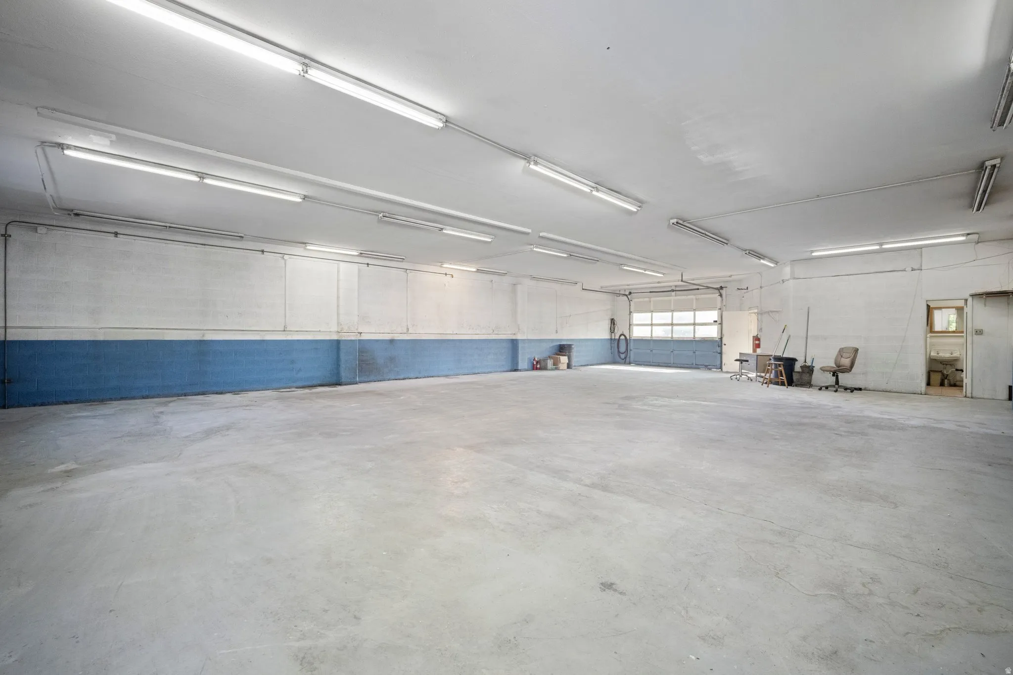 Expansive concrete flooring with high ceilings, featuring fluorescent lighting fixtures, a large overhead door, and a utility sink