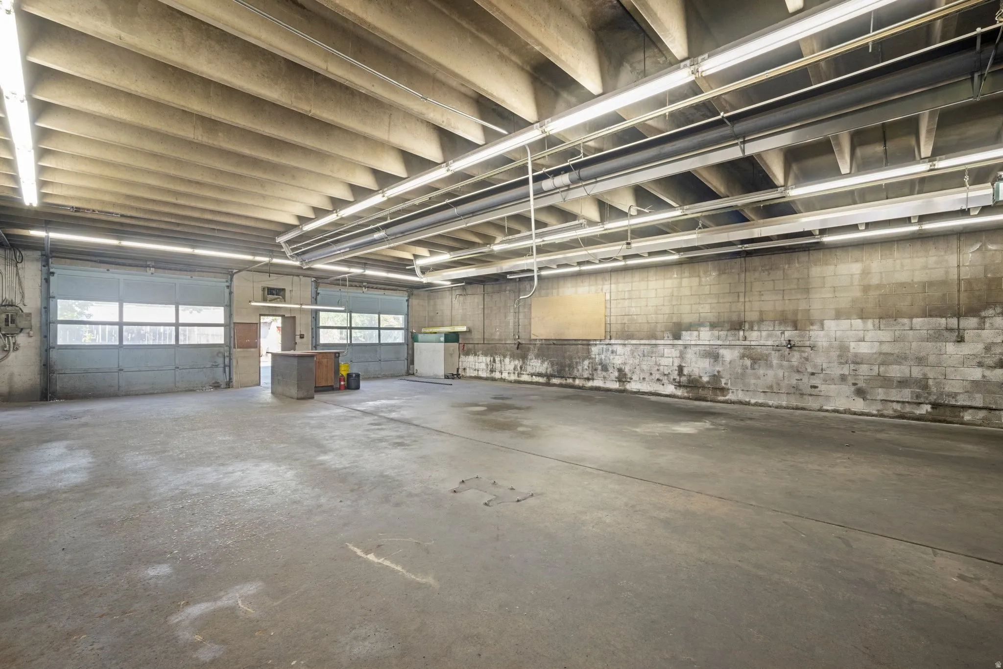 Concrete slab flooring, two overhead roll-up doors with windows, exposed concrete block walls, overhead fluorescent lighting, and exposed ceiling joists