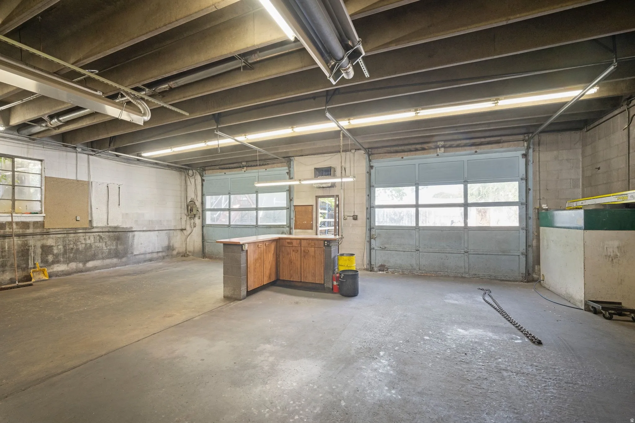 Open floor plan with concrete flooring, multiple overhead fluorescent light fixtures, exposed ceiling beams, and built-in wood-finish cabinetry