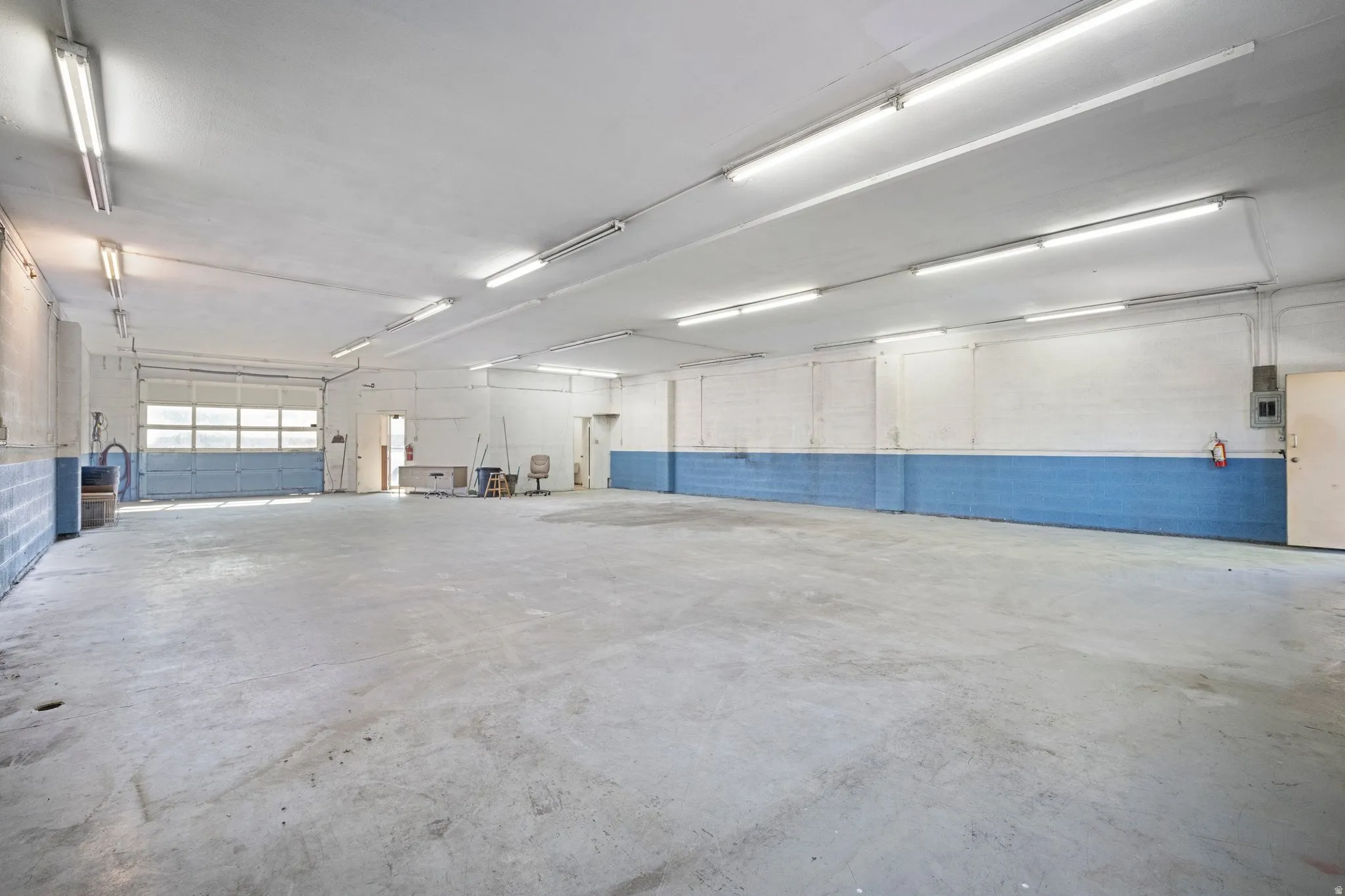 Spacious garage with concrete flooring, multiple fluorescent lighting fixtures, a large roll-up door, and block wall construction