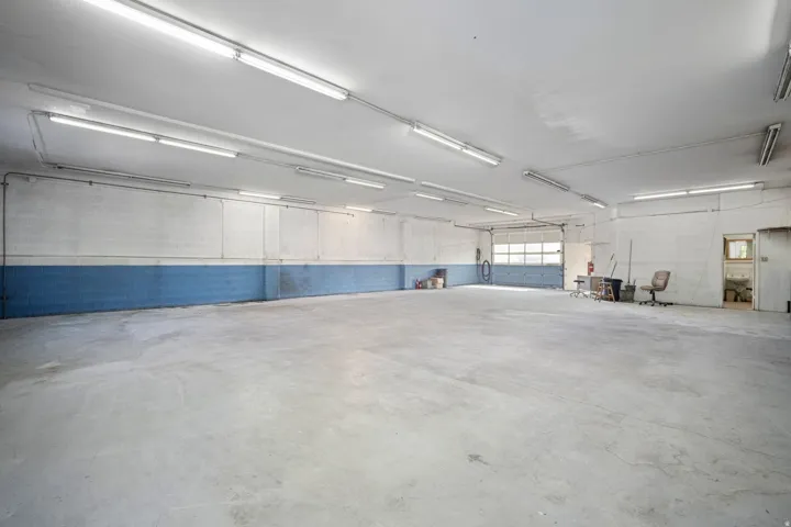 Expansive concrete flooring with high ceilings, featuring fluorescent lighting fixtures, a large overhead door, and a utility sink