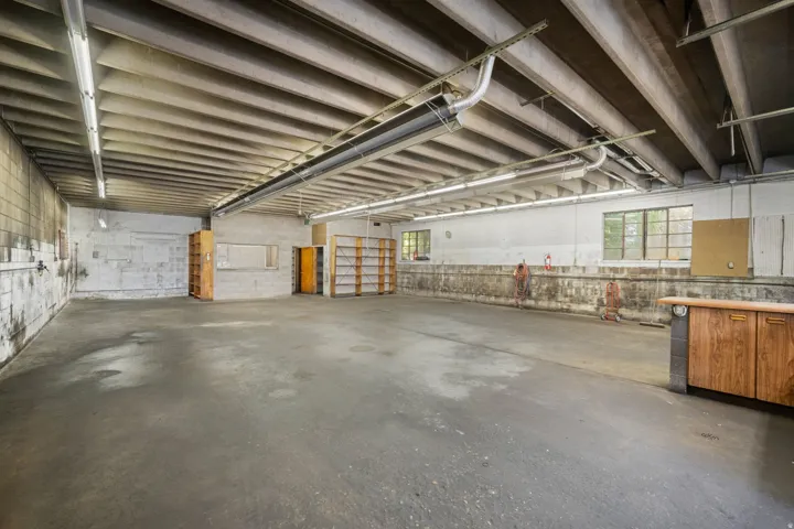 Expansive concrete flooring, exposed ceiling beams, built-in shelving, multiple windows, and fluorescent overhead lighting
