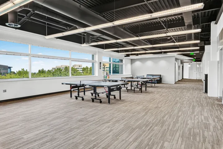Expansive open-plan area featuring large windows, patterned carpet flooring, exposed ceiling, and linear fluorescent lighting