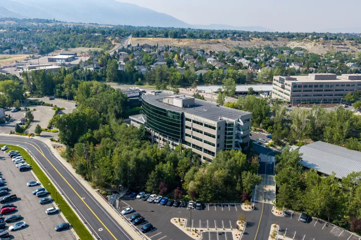 Modern office building featuring a curved glass facade, extensive parking areas, surrounding mature trees, and a mountain backdrop