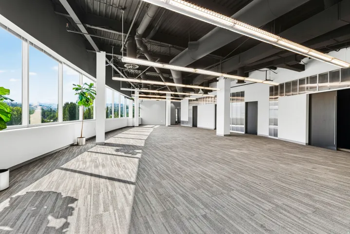 Expansive commercial space featuring a full wall of tall windows, exposed ductwork ceiling, suspended linear lighting, and patterned carpet flooring