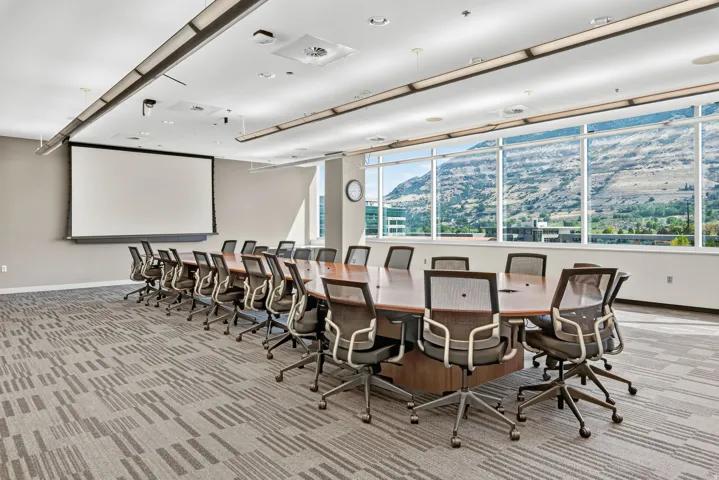 Large conference room with a substantial wood-finish table, a wall-mounted projection screen, and integrated ceiling lighting