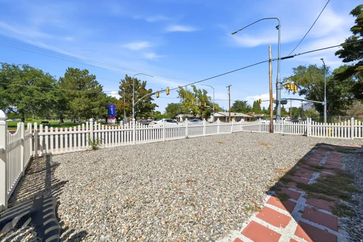 Spacious gravel-surfaced yard enclosed by white picket fencing, mature trees providing natural shade, overhead utility lines and street lighting, with paved red accent edging along the perimeter