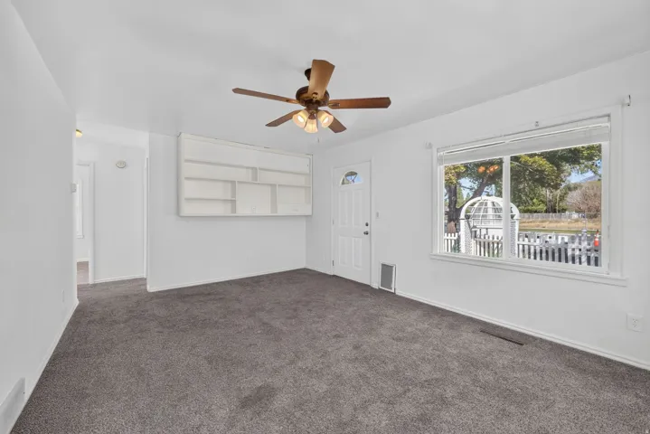 Recessed built-in shelving, white paneled entry door, flush-mounted ceiling fan, large window with white frame, and gray carpeting