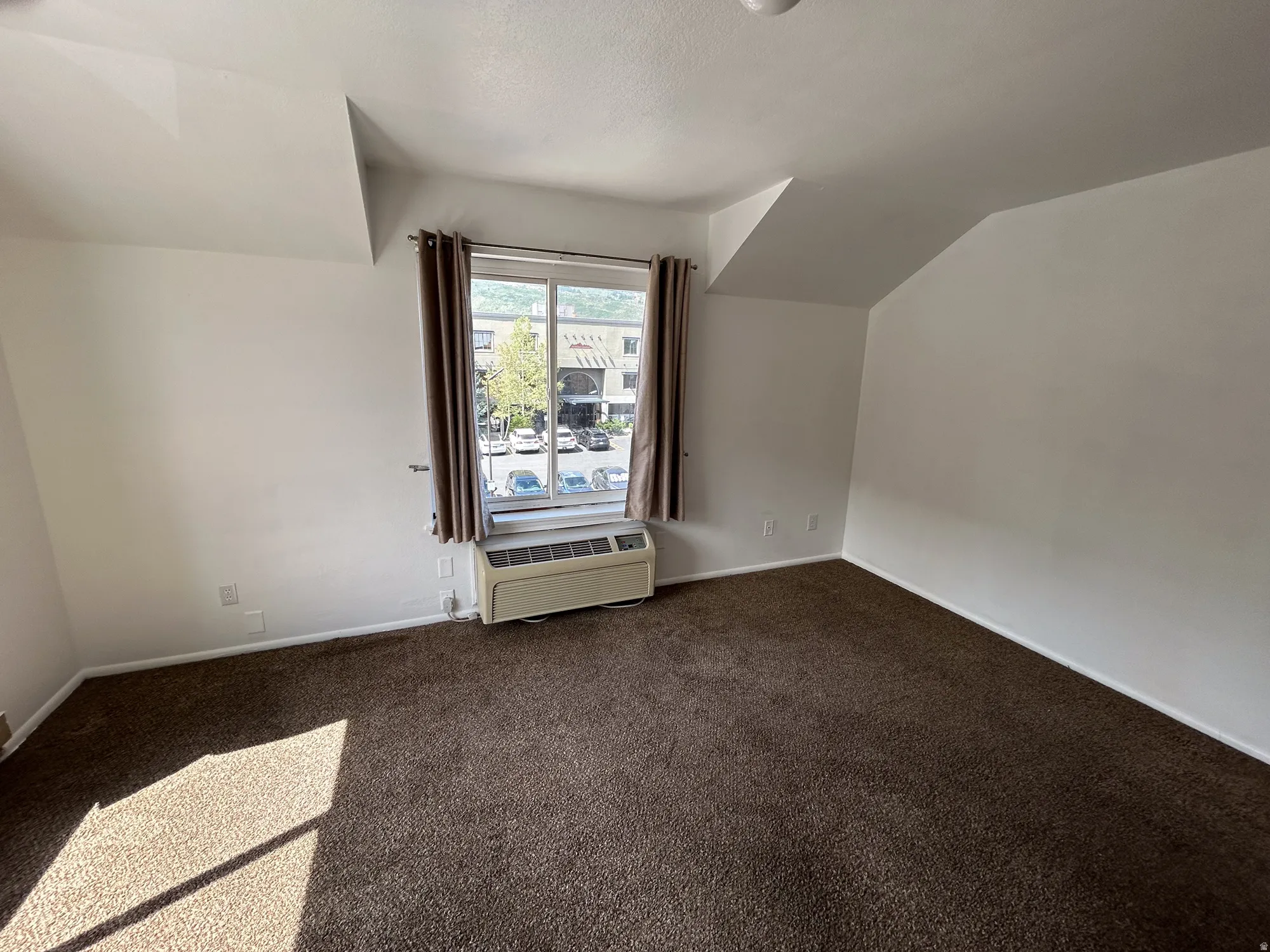 Sloped ceiling room featuring brown carpet flooring and a window with a compact air conditioning unit