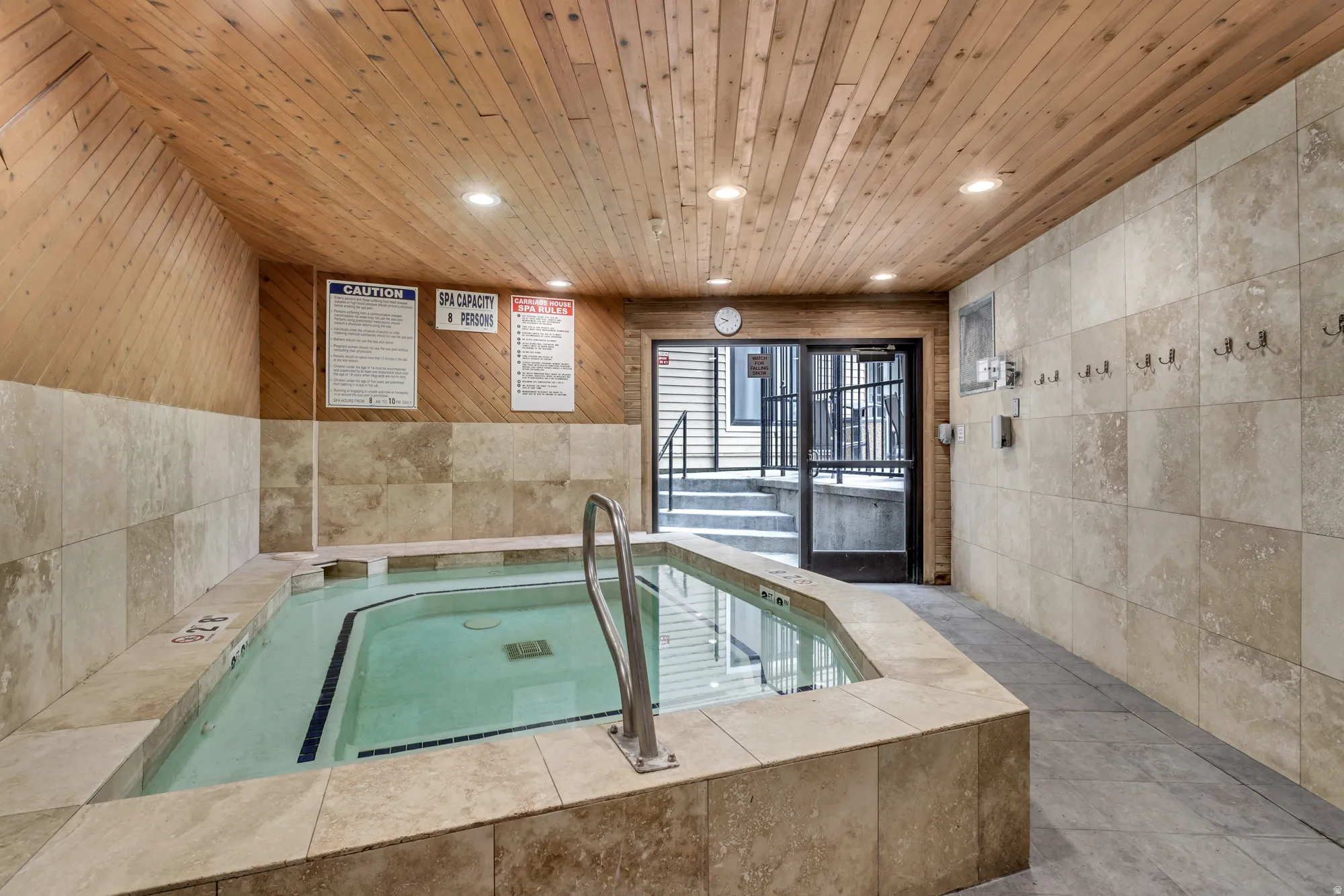 Spa-inspired indoor soaking tub with tile surround and integrated handrail, complemented by a wood-finish ceiling with recessed lighting, neutral stone-finish wall tiles, and direct access to an outdoor patio area through glass doors