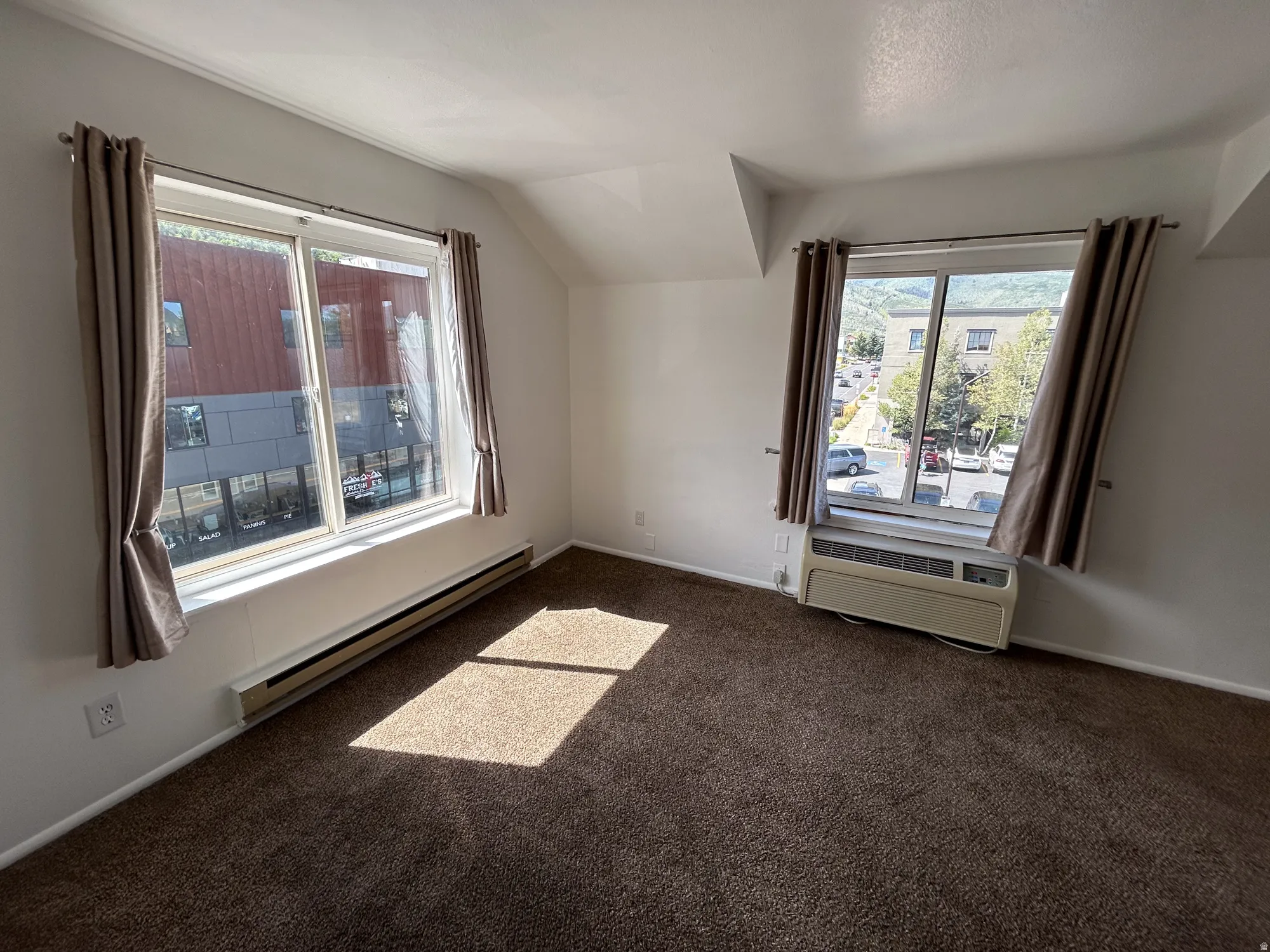 Carpeted room with multiple windows, featuring baseboard heating and a wall-mounted air conditioning unit