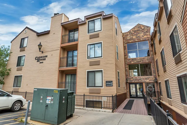 Multi-story building featuring tan horizontal siding, stone facade accents, multiple private balconies with black railings, and a prominent entrance with double glass doors