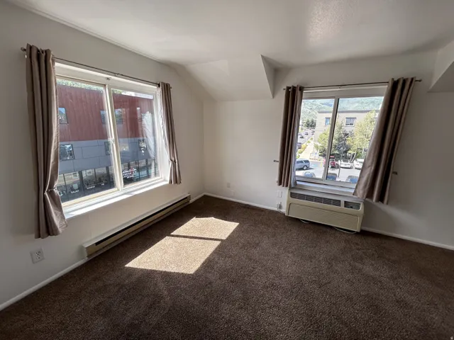 Carpeted room with multiple windows, featuring baseboard heating and a wall-mounted air conditioning unit