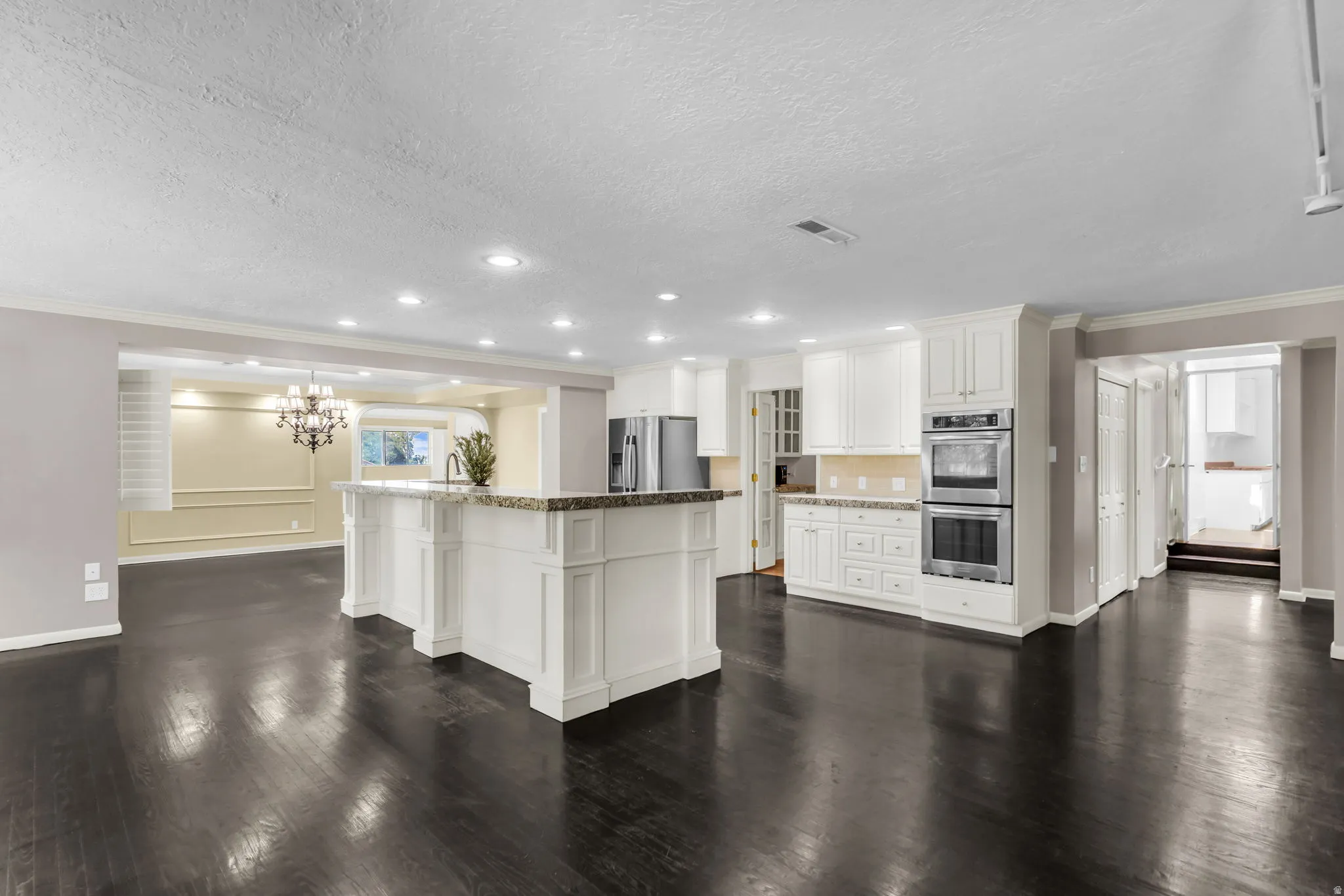 Spacious kitchen featuring white cabinetry, stone countertops, a large island with seating, dark wood-finish flooring, and built-in stainless steel double ovens