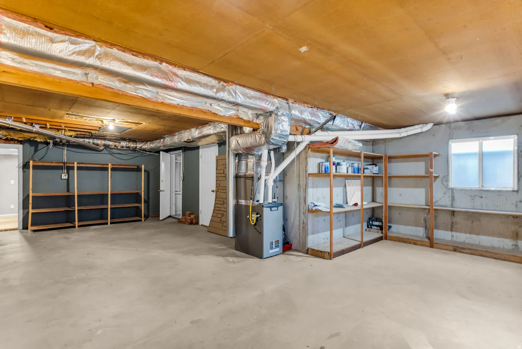 Unfinished basement with concrete flooring, exposed ceiling beams, built-in shelving, and a window