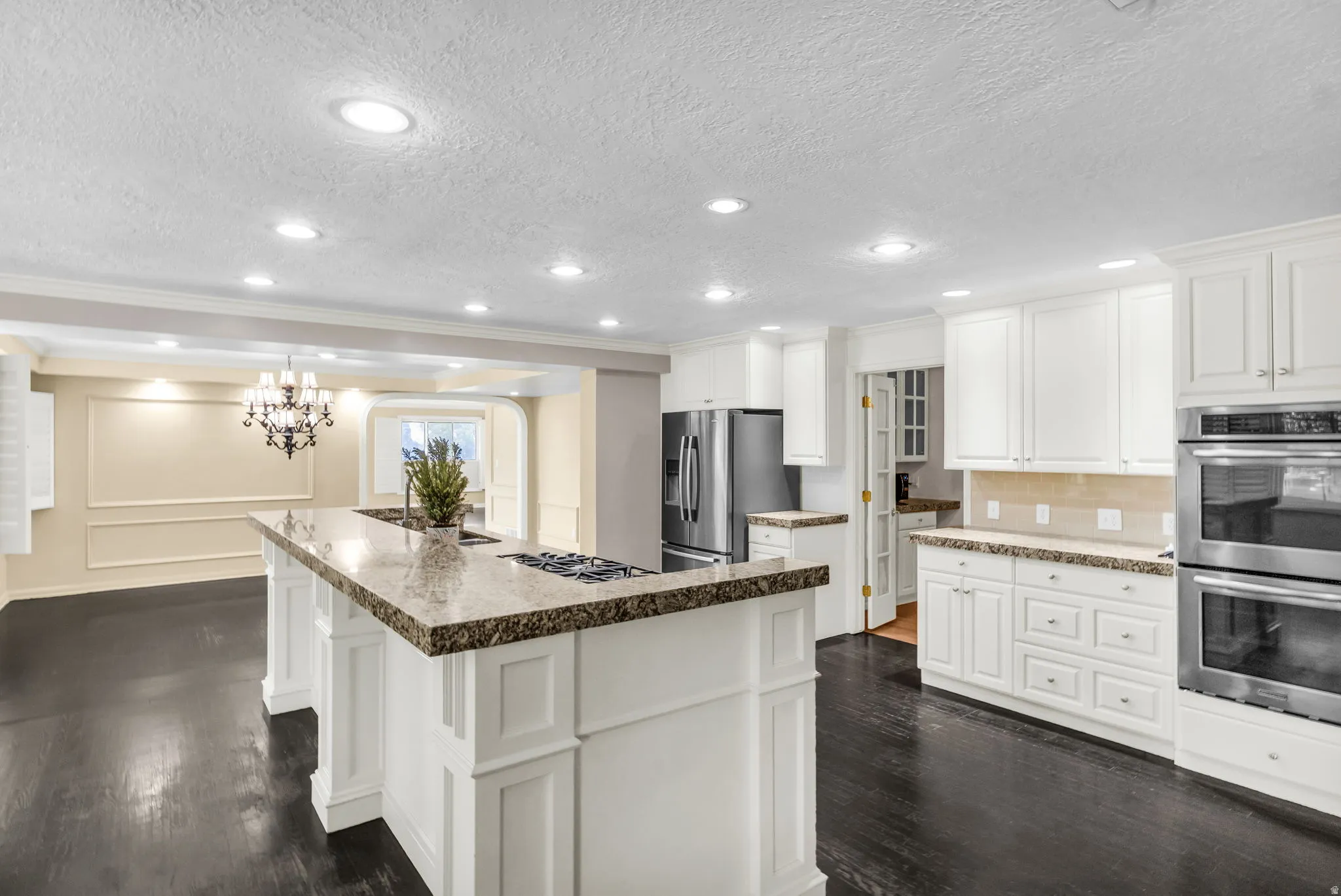 Gourmet kitchen featuring a large center island with decorative base panels and stone-top countertop, white cabinetry with raised-panel doors, stainless steel appliances including dual wall ovens, recessed ceiling lighting, dark hardwood flooring, and a b
