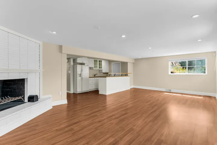 Spacious living area featuring wood-finish flooring, a white brick fireplace with a dark hearth, recessed lighting, and a window providing natural illumination