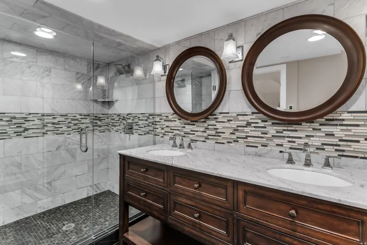 Dual vanity with wood cabinetry and undermount sinks, stone countertop, two framed mirrors, glass-enclosed shower with tiled walls, and mosaic tile accent