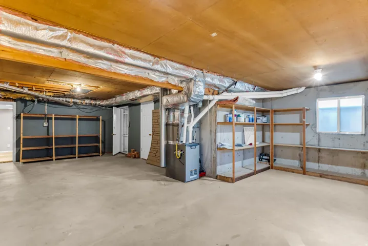 Unfinished basement with concrete flooring, exposed ceiling beams, built-in shelving, and a window