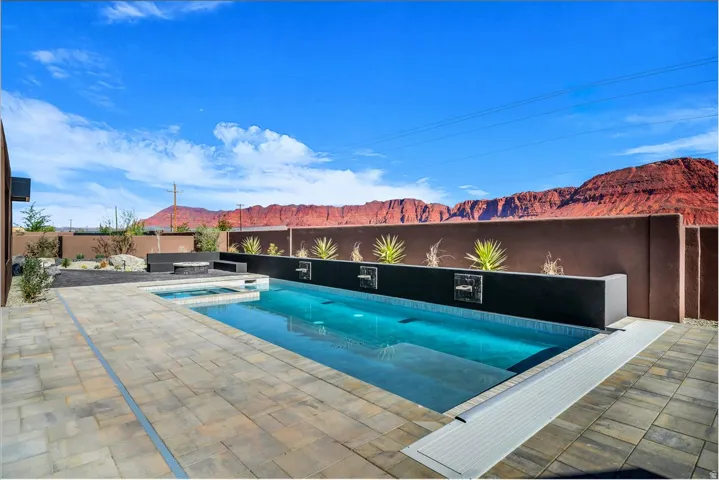 Paver stone patio surrounding a swimming pool with a built-in spa, dark accent wall with water features, and a fire pit