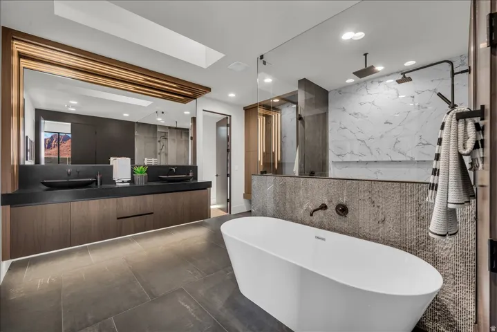 Freestanding soaking tub with dark finish fixtures, dual vanity with undermount lighting, dark wood-finish cabinetry, and a large format mirror