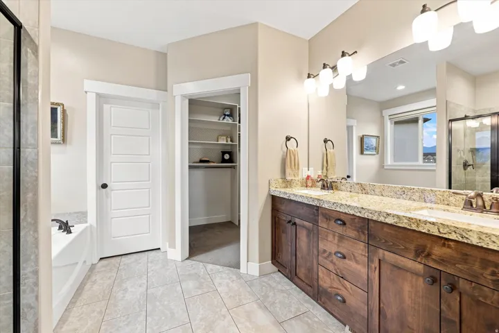 Dual vanity with granite countertop, wood-finish cabinetry, tile flooring, built-in bathtub, and walk-in shower with glass enclosure