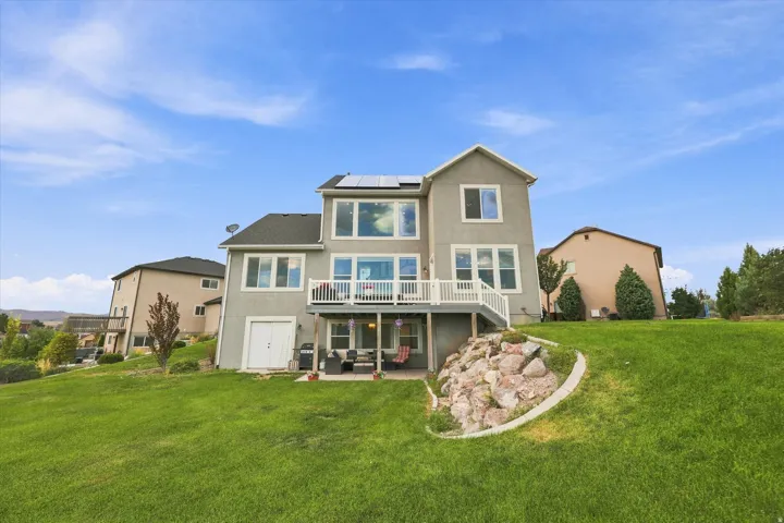 Expansive green lawn, two-story stucco exterior, upper-level deck with white railing, lower-level patio, and sloped landscaping with rock features