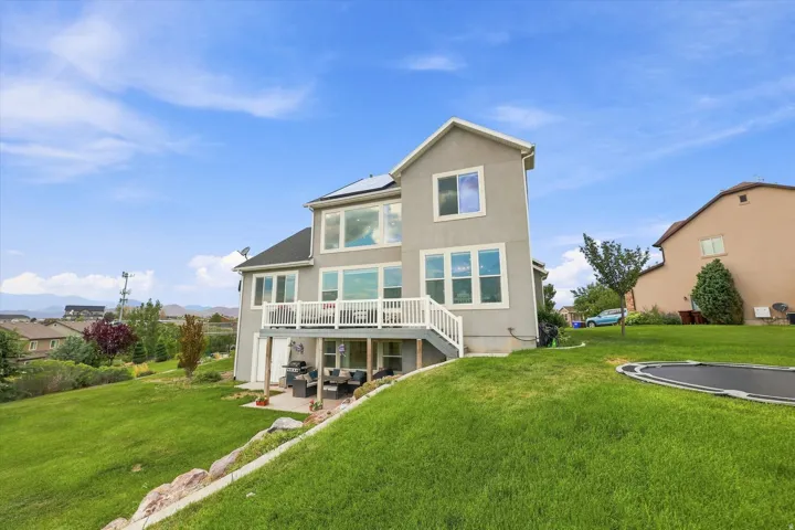 Two-story stucco exterior with multiple windows, upper-level deck, ground-level patio, and sloped grassy terrain