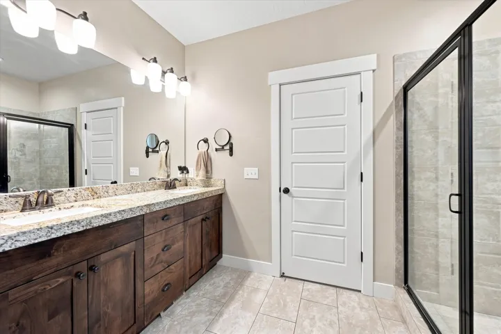 Dual vanity with stone countertop, dark wood-finish cabinetry, tile flooring, and a glass-enclosed shower
