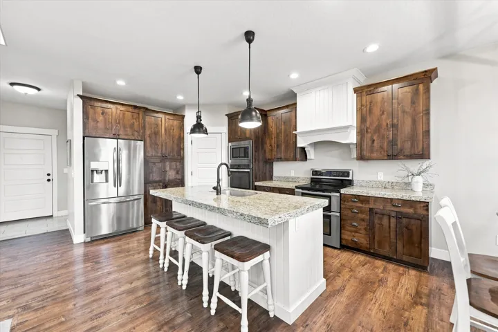 Spacious kitchen featuring wood-finish flooring, dark wood cabinetry, stainless steel appliances, a white beadboard island with a granite-style countertop, and pendant lighting